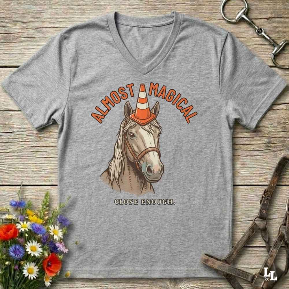 A heather gray V-neck t-shirt laid flat on a rustic wooden surface, featuring a graphic of a horse wearing an orange and white traffic cone hat, with the text 'ALMOST MAGICAL' above and 'CLOSE ENOUGH.' below.