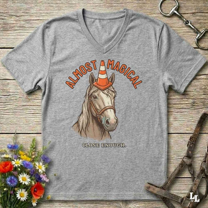 A heather gray V-neck t-shirt laid flat on a rustic wooden surface, featuring a graphic of a horse wearing an orange and white traffic cone hat, with the text 'ALMOST MAGICAL' above and 'CLOSE ENOUGH.' below.