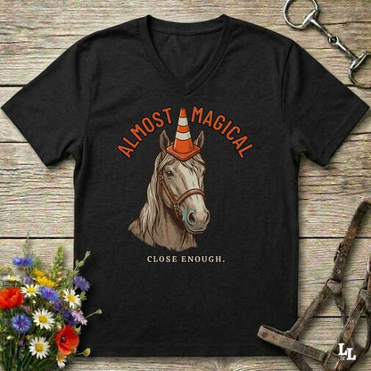 A black V-neck t-shirt laid flat on a rustic wooden surface, featuring a graphic of a horse wearing an orange-and-white traffic cone with the words 'ALMOST MAGICAL' above and 'CLOSE ENOUGH.' below.