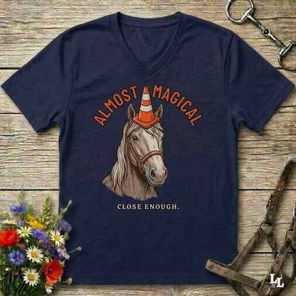A navy blue V-neck t-shirt laid flat on a rustic wooden surface, featuring a graphic of a horse with a traffic cone on its horn and the text 'ALMOST MAGICAL' and 'CLOSE ENOUGH.' Decorative flowers and equestrian gear are placed around the shirt.