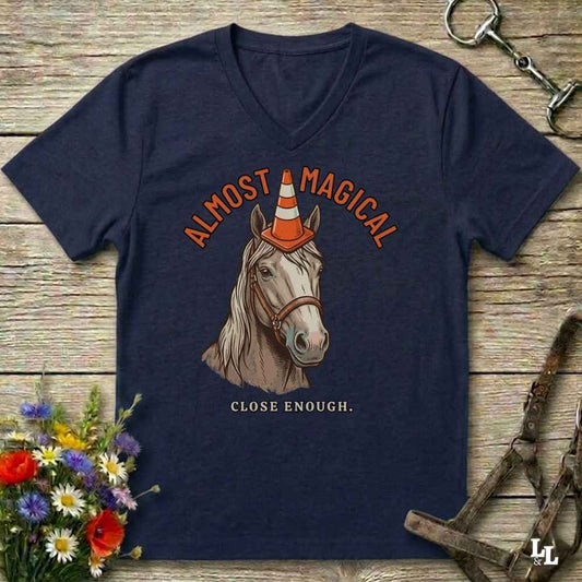 A navy blue V-neck t-shirt laid flat on a rustic wooden surface, featuring a graphic of a horse with a traffic cone on its horn and the text 'ALMOST MAGICAL' and 'CLOSE ENOUGH.' Decorative flowers and equestrian gear are placed around the shirt.