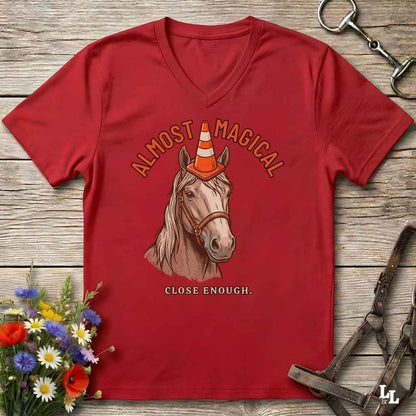 A red V-neck t-shirt laid flat on a rustic wooden surface, featuring a graphic of a horse wearing an orange and white traffic cone with the text 'ALMOST MAGICAL' above and 'CLOSE ENOUGH.' below.