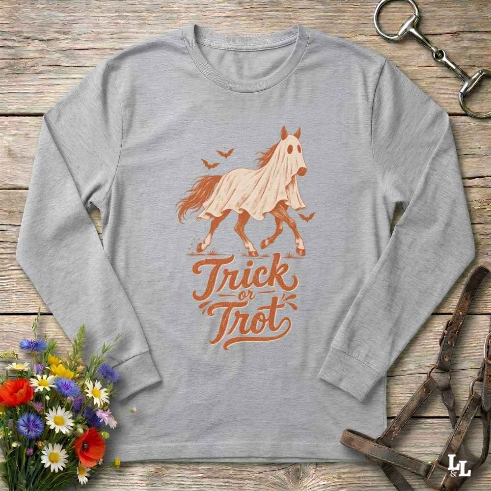 A heather gray long-sleeve t-shirt with a graphic of a ghosted horse and the words 'Trick or Treat' in orange-brown lettering, laid flat on a rustic wooden surface with decorative flowers and horse tack around it.