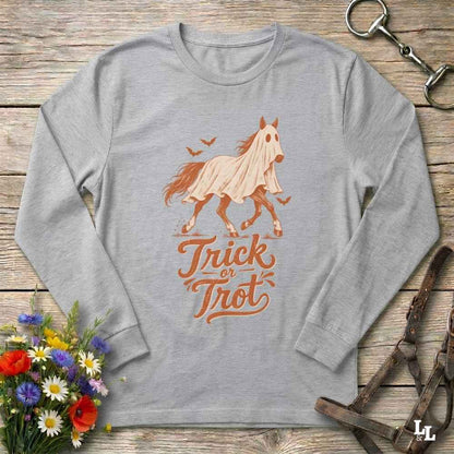A heather gray long-sleeve t-shirt with a graphic of a ghosted horse and the words 'Trick or Treat' in orange-brown lettering, laid flat on a rustic wooden surface with decorative flowers and horse tack around it.