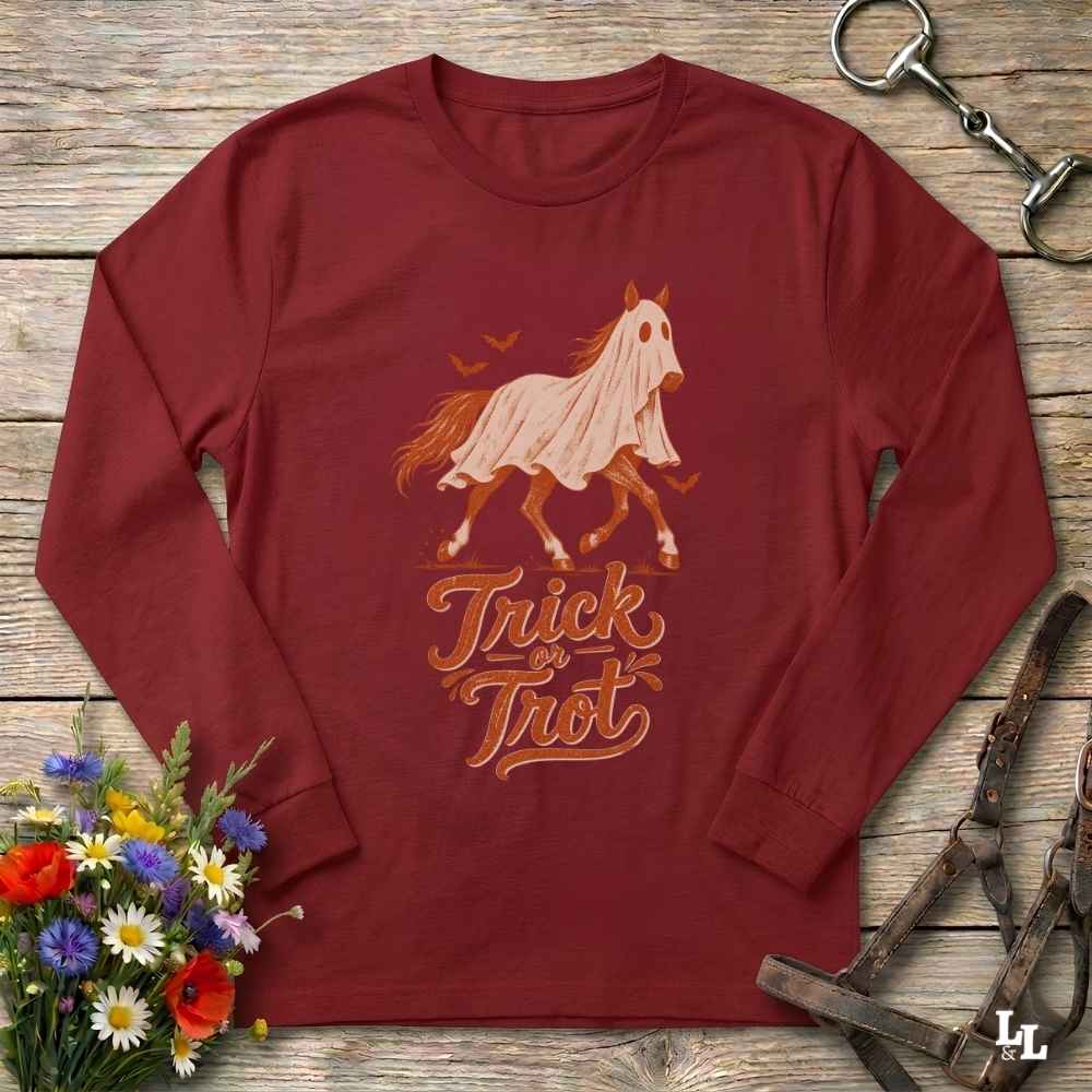 A long-sleeve crewneck shirt in burgundy red, featuring a graphic of a ghosted horse in a white sheet with the words 'Trick or Treat' underneath. The shirt is displayed in a flat-lay arrangement on a rustic wooden background, surrounded by equestrian tack and a small bouquet of wildflowers.
