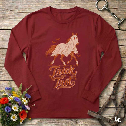 A long-sleeve crewneck shirt in burgundy red, featuring a graphic of a ghosted horse in a white sheet with the words 'Trick or Treat' underneath. The shirt is displayed in a flat-lay arrangement on a rustic wooden background, surrounded by equestrian tack and a small bouquet of wildflowers.