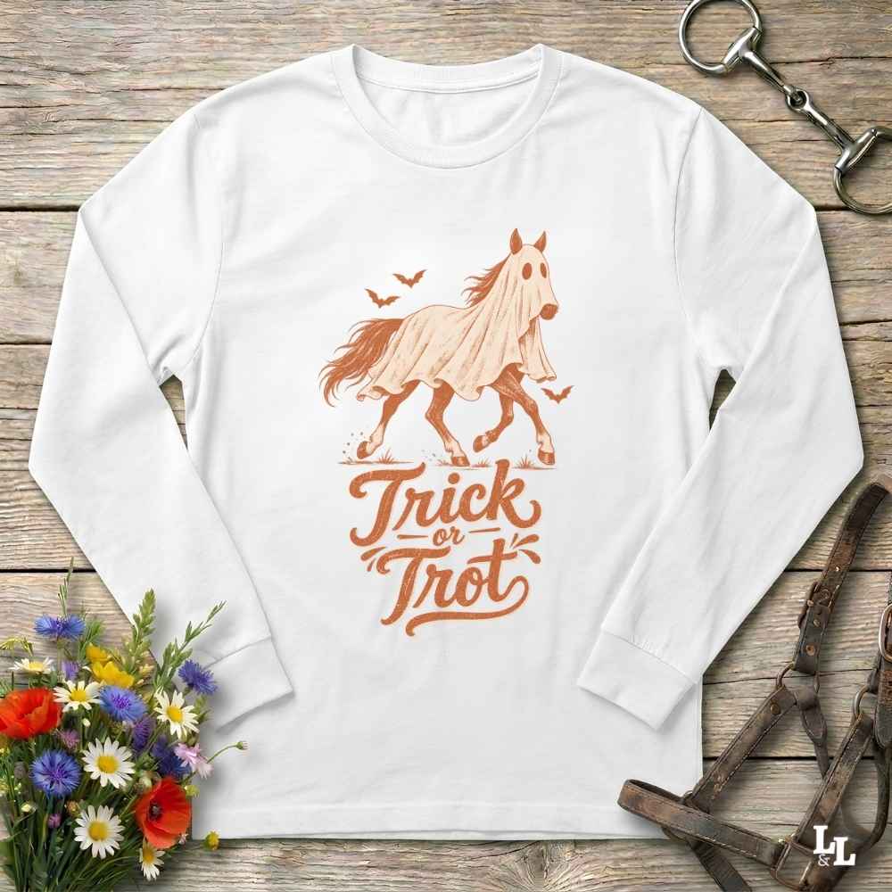 A white long-sleeve t-shirt with a 'Trick or Trot' horse graphic, laid flat on a rustic wooden background with equestrian accessories and flowers.