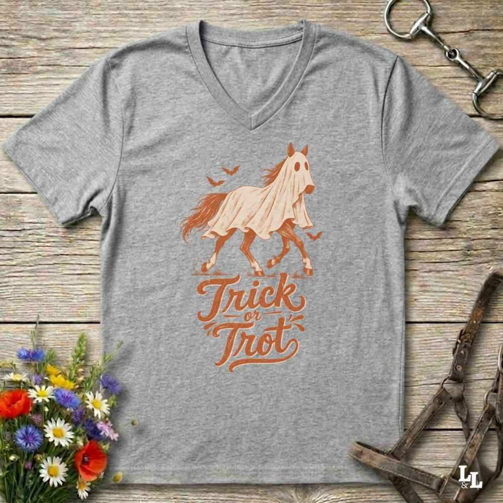 A heather gray V-neck t-shirt featuring a graphic of a ghostly horse in a sheet with the text 'Trick or Treat' in orange. The shirt is displayed flat on a rustic wooden background with equestrian and floral props.