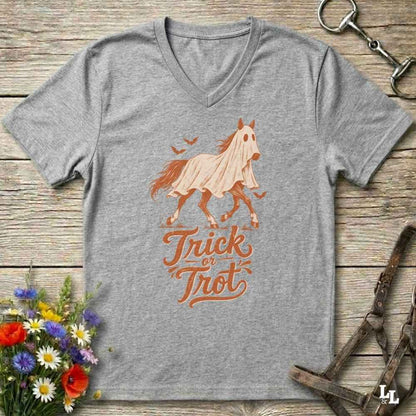 A heather gray V-neck t-shirt featuring a graphic of a ghostly horse in a sheet with the text 'Trick or Treat' in orange. The shirt is displayed flat on a rustic wooden background with equestrian and floral props.