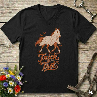 A black V-neck t-shirt featuring a graphic of a white horse with an orange mane and the words 'Trick or Treat' in orange script, laid flat on a rustic wooden background with equestrian and floral props.