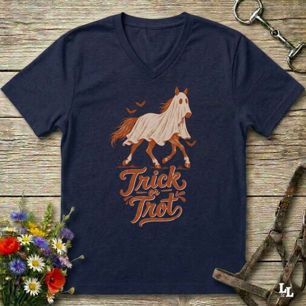 A navy blue V-neck t-shirt laid flat on a rustic wooden surface, featuring a graphic of a ghostly horse in a white sheet with the text 'Trick or Treat' in orange-brown. The shirt is styled with equestrian tack in the background and a small bouquet of wildflowers in the lower-left corner.