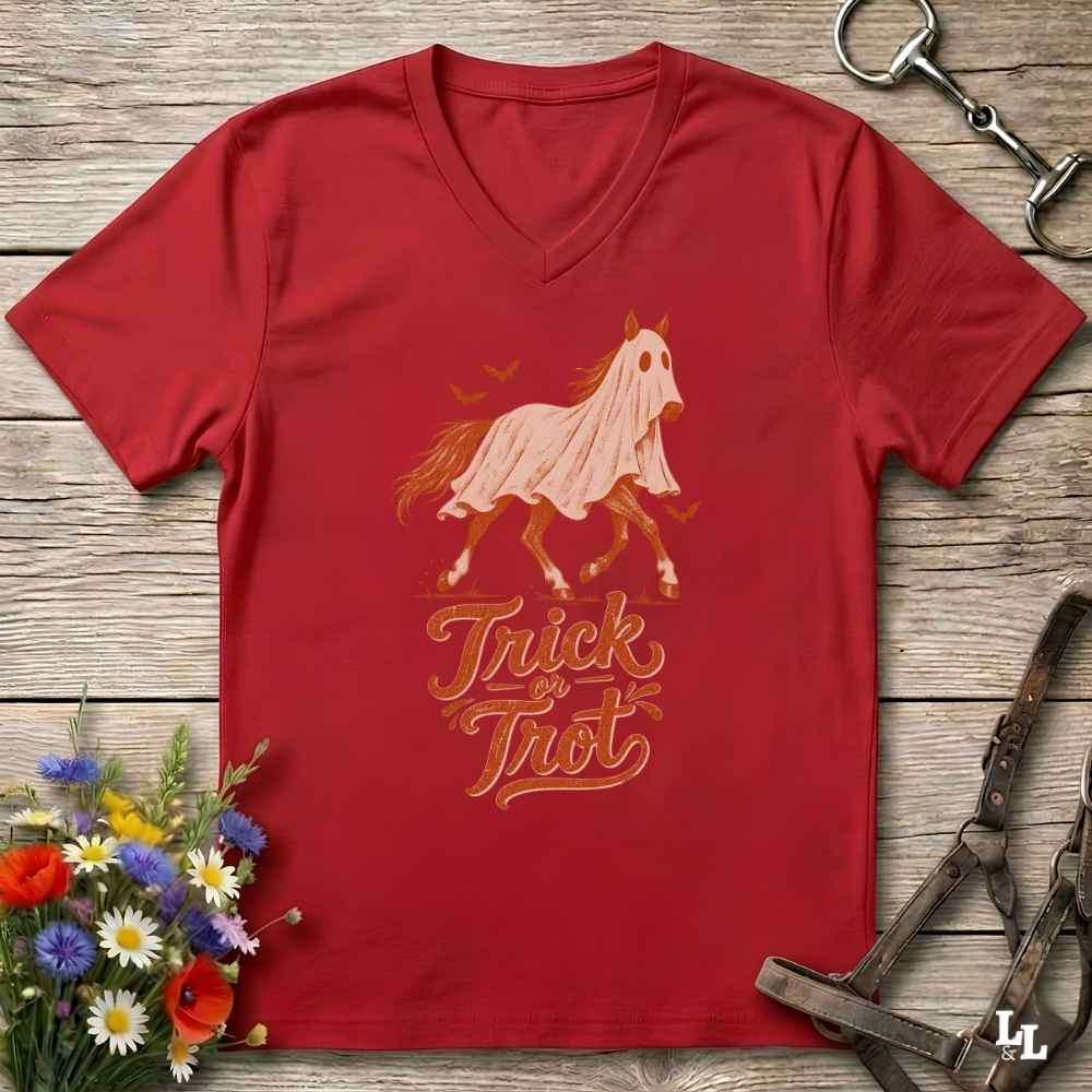 A red V-neck t-shirt laid flat on a rustic wood background, featuring a graphic of a horse draped in a white sheet with the words 'Trick or Treat' in orange. The shirt is styled with equestrian tack and a small bouquet of flowers.