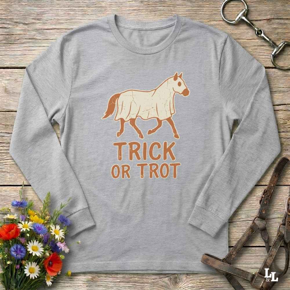 A light gray long-sleeve t-shirt with a graphic of a horse wearing a white blanket and the text 'TRICK OR TROT' in brown. The shirt is displayed in a flat-lay arrangement on a rustic wooden background, surrounded by decorative props including a bouquet of flowers, a horse bit, and a leather halter.