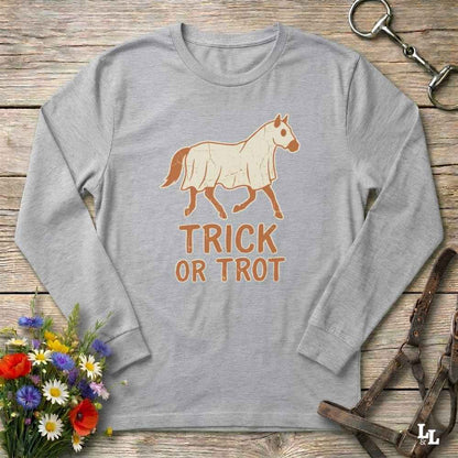 A light gray long-sleeve t-shirt with a graphic of a horse wearing a white blanket and the text 'TRICK OR TROT' in brown. The shirt is displayed in a flat-lay arrangement on a rustic wooden background, surrounded by decorative props including a bouquet of flowers, a horse bit, and a leather halter.
