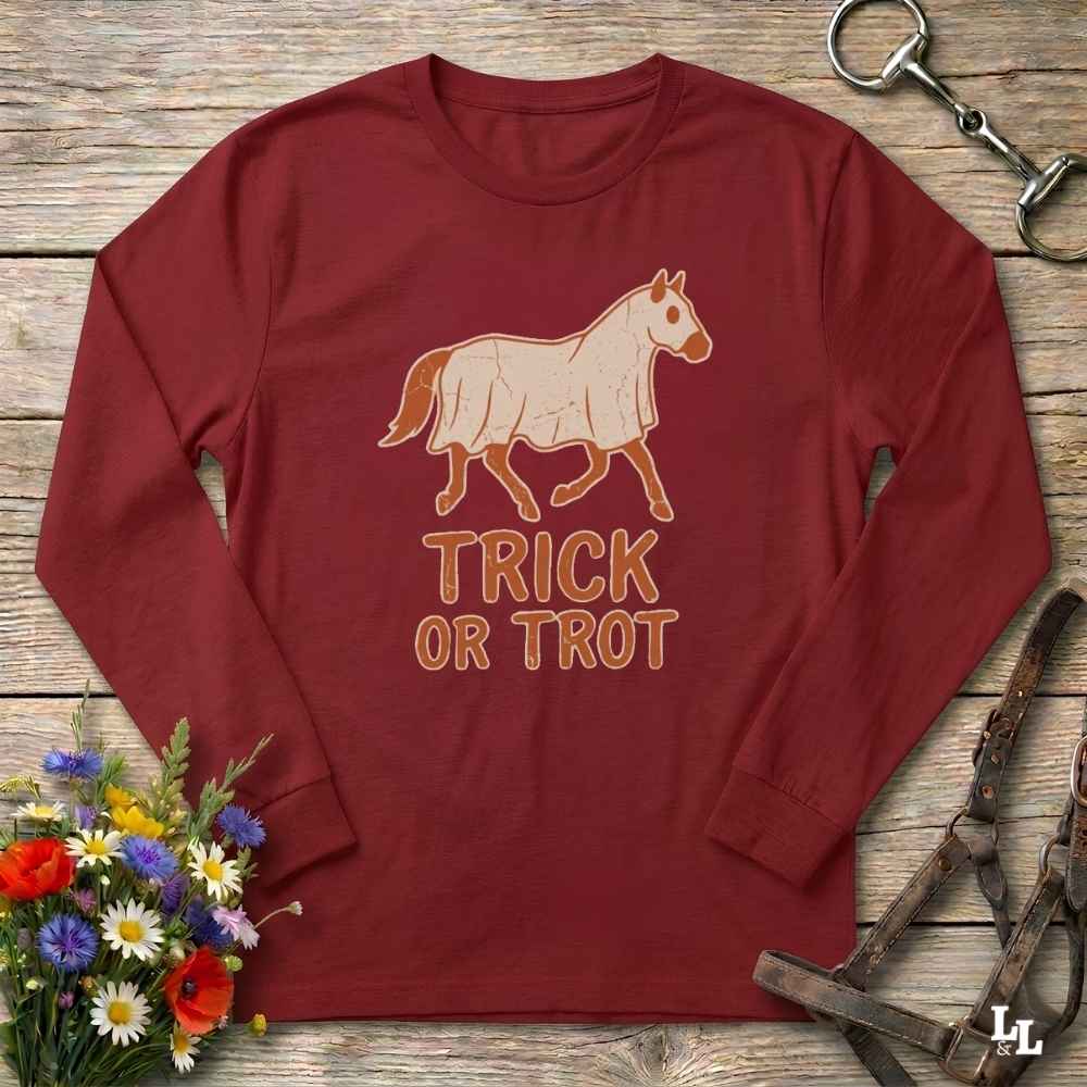 A long-sleeve crewneck t-shirt in maroon red, laid flat on a rustic wooden background. The shirt features a graphic of a horse with a cutaway torso showing internal organs, above the text 'TRICK OR TROT' in a matching outline font.