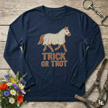 A navy blue long-sleeve t-shirt with a graphic of a horse wearing a white blanket and the text 'TRICK OR TROT' in brown. The shirt is displayed flat on a rustic wooden background with equestrian accessories and a small flower arrangement.