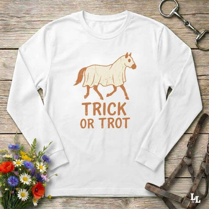 A white long-sleeve t-shirt with a graphic of a horse in a coat and the text 'TRICK OR TROT' in orange, laid flat on a rustic wooden background with decorative flowers and tack.