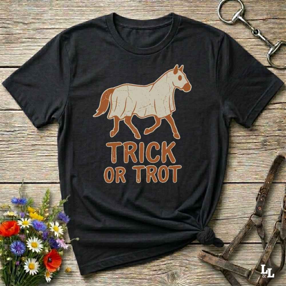 A black short-sleeve graphic T-shirt laid flat on a rustic wood surface, featuring a tan horse illustration and the words “TRICK OR TROT” in tan lettering. Decorative flowers and horse tack are placed around the shirt.