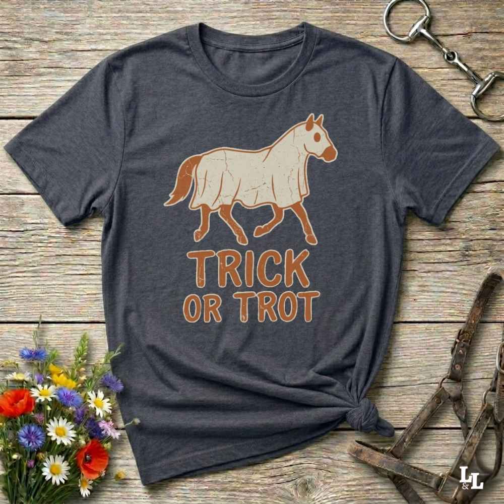 A dark heather gray t-shirt with a graphic of a white horse and the text 'TRICK OR TROT' in orange-brown, laid flat on a rustic wooden surface with decorative flowers and horse tack.