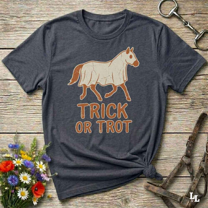 A dark heather gray t-shirt with a graphic of a white horse and the text 'TRICK OR TROT' in orange-brown, laid flat on a rustic wooden surface with decorative flowers and horse tack.