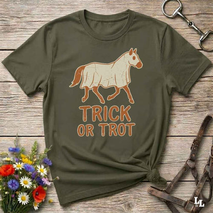 An olive green short-sleeve t-shirt with a graphic of a white horse and the text 'TRICK OR TROT' in orange-brown, laid flat on a rustic wooden surface with decorative flowers and horse tack.