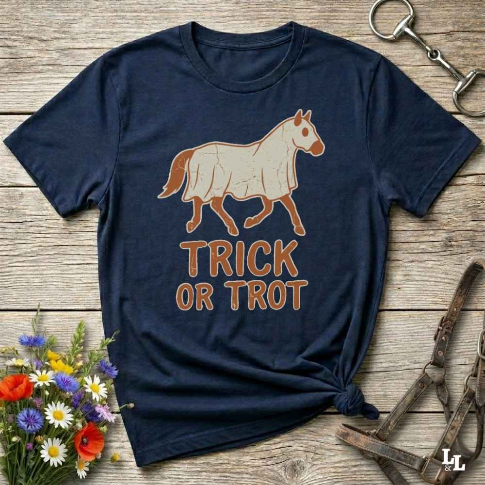 A navy blue graphic t-shirt featuring a cream-colored horse illustration and the words 'TRICK OR TROT' in brown, styled on a rustic wooden background with equestrian accessories and flowers.