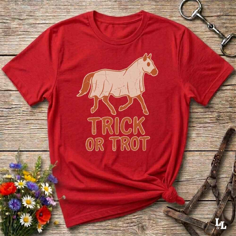 A red short-sleeve graphic T-shirt laid flat on a rustic wooden surface, with a tan horse illustration and the words 'TRICK OR TROT' printed on the front. The shirt is styled with flowers, a halter, and a metal ring around it.