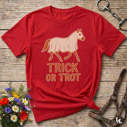 A red short-sleeve graphic T-shirt laid flat on a rustic wooden surface, with a tan horse illustration and the words 'TRICK OR TROT' printed on the front. The shirt is styled with flowers, a halter, and a metal ring around it.