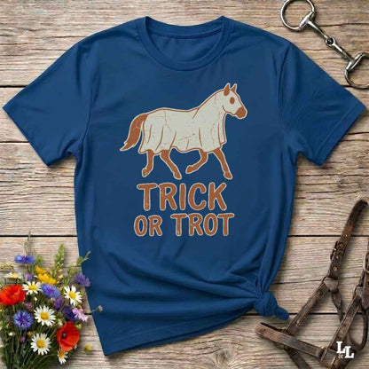 A navy blue graphic t-shirt laid flat on a rustic wooden surface, featuring a cream-colored horse illustration and the words 'TRICK OR TROT' in brown. The shirt is styled with a small bouquet of wildflowers on the bottom left and horse tack on the right.