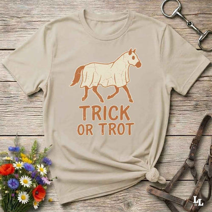 A beige graphic t-shirt with a horse illustration and the text 'TRICK OR TROT' printed in orange, styled in a flat-lay on a rustic wooden background with equestrian accessories and flowers.