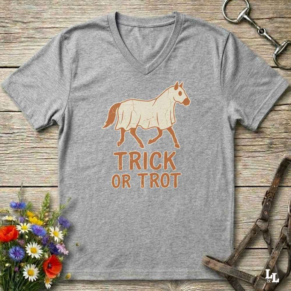 A heather gray V-neck t-shirt featuring a graphic of a horse with a harness and the text 'TRICK OR TROT' in orange-brown. The shirt is displayed flat on a rustic wooden background with equestrian-themed props like a horse bit, bridle, and a small bouquet of wildflowers.