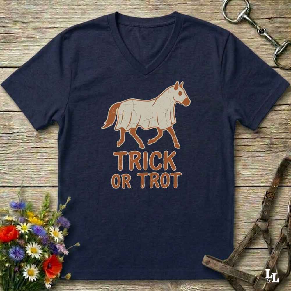 A navy blue V-neck t-shirt with a graphic of a white and brown horse and the text 'TRICK OR TROT' in brown, laid flat on a rustic wooden background with equestrian accessories and flowers.
