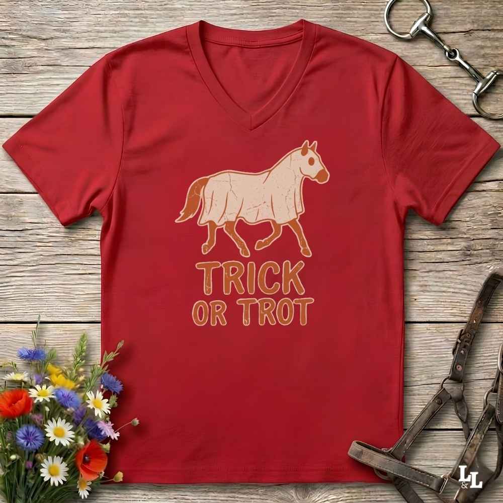 A red V-neck t-shirt laid flat on a rustic wooden surface, featuring a graphic of a horse wearing a blanket with the text 'TRICK OR TROT' underneath. The shirt is styled with equestrian accessories (a bit and bridle) and a small bouquet of wildflowers.