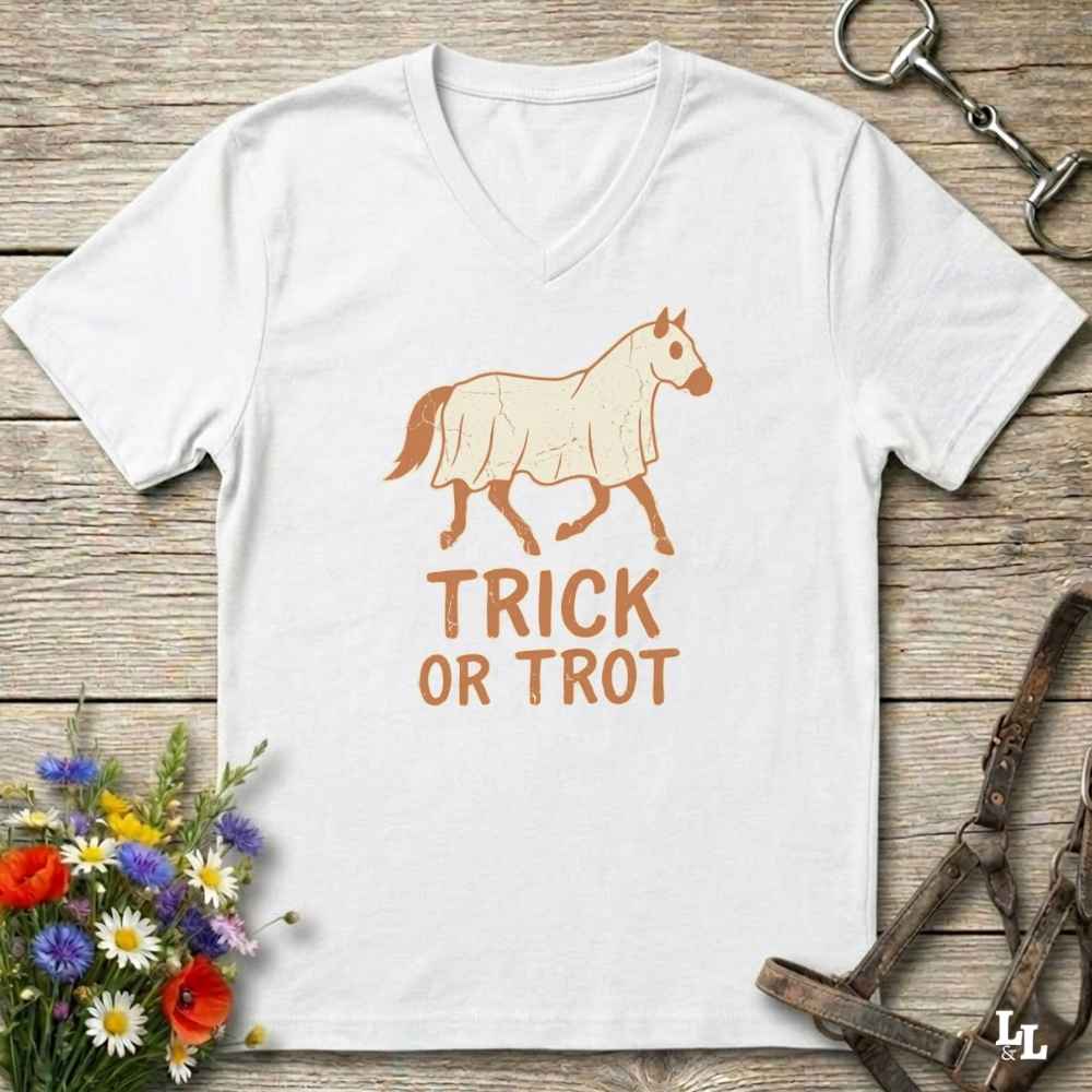 A white v-neck t-shirt with a graphic of a horse wearing a coat and the text 'TRICK OR TROT' in orange-brown, displayed on a rustic wooden background with equestrian accessories and flowers.