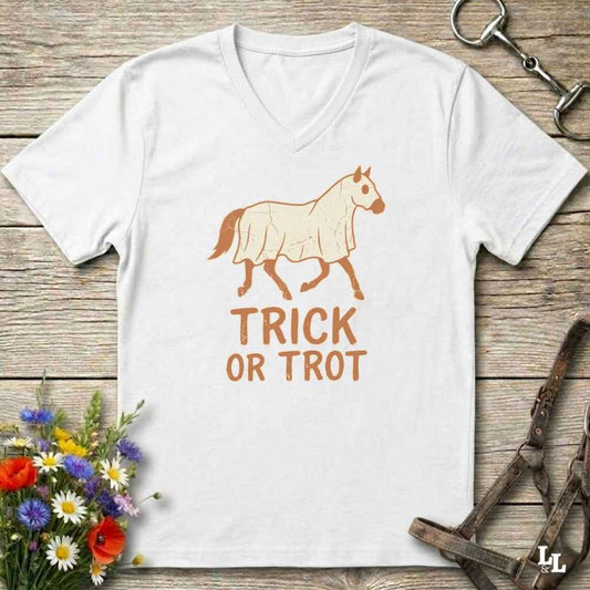 A white v-neck t-shirt with a graphic of a horse wearing a coat and the text 'TRICK OR TROT' in orange-brown, displayed on a rustic wooden background with equestrian accessories and flowers.
