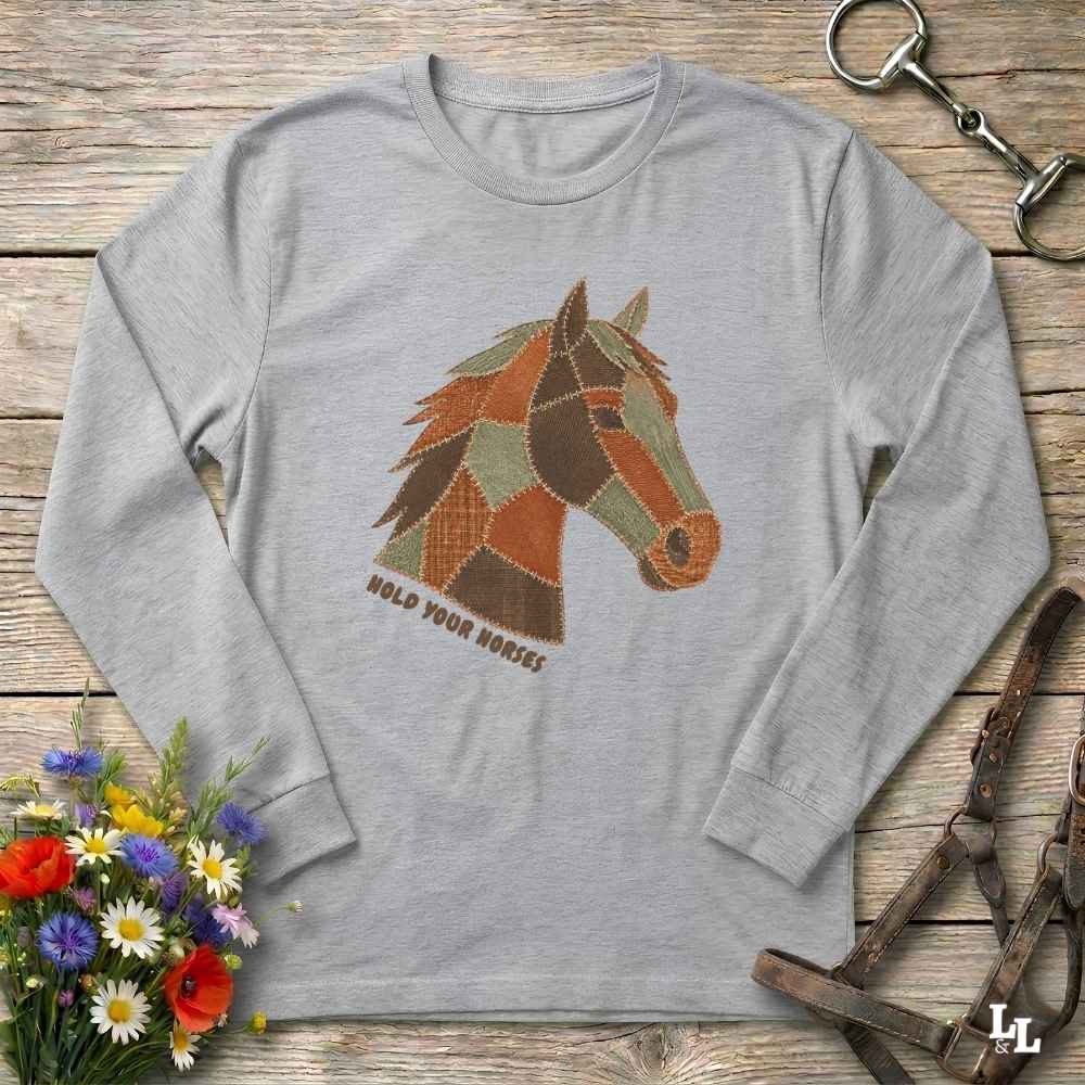 A light gray long-sleeve t-shirt with a geometric horse head graphic in earthy tones and the text 'HOLD YOUR HORSES' printed below it. The shirt is displayed flat on a rustic wooden background with equestrian-themed props including a halter, tack, and a small bouquet of wildflowers.