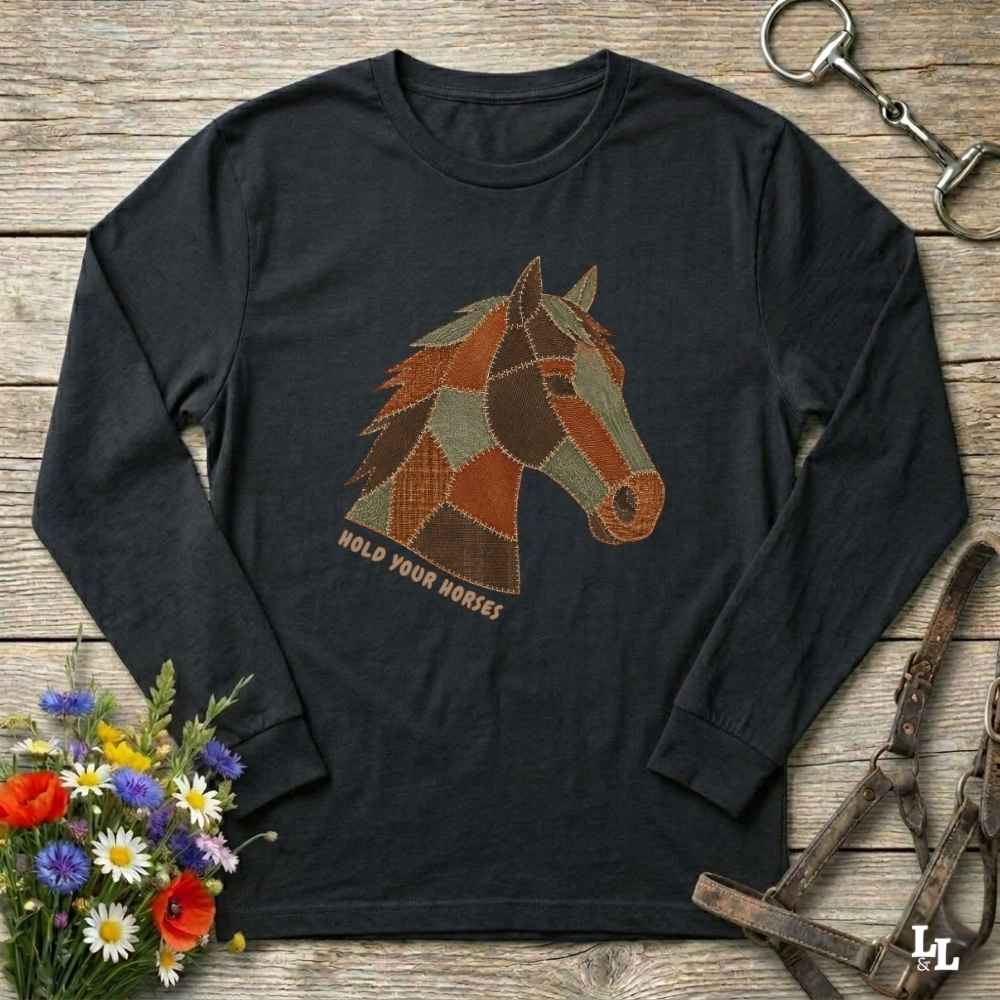 A black long-sleeve t-shirt with a geometric patchwork horse head graphic in brown, orange, and beige, with the text 'HOLD YOUR HORSES' printed below it. The shirt is displayed flat on a rustic wooden background with equestrian-themed props including a halter, a horse bit, and a small bouquet of wildflowers.