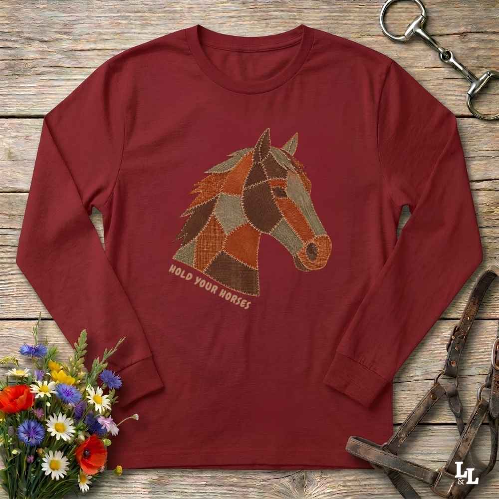 A long-sleeve crewneck t-shirt in maroon red, featuring a graphic of a horse head made of patchwork shapes in brown, orange, and beige, with the text 'HOLD YOUR HORSES' printed below. The shirt is displayed flat on a rustic wooden background with equestrian-themed props including a halter, reins, and a small bouquet of wildflowers.