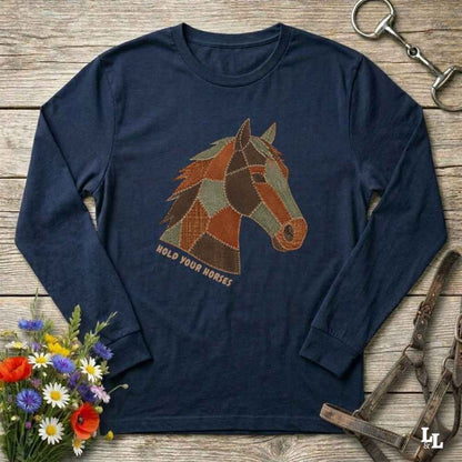 A navy blue long-sleeve t-shirt with a patchwork-style horse head graphic and the text 'HOLD YOUR HORSES' printed on the front. The shirt is displayed in a flat-lay arrangement on a rustic wooden background, surrounded by equestrian-themed accessories (a metal horse bit, leather bridle, and a small bouquet of wildflowers).