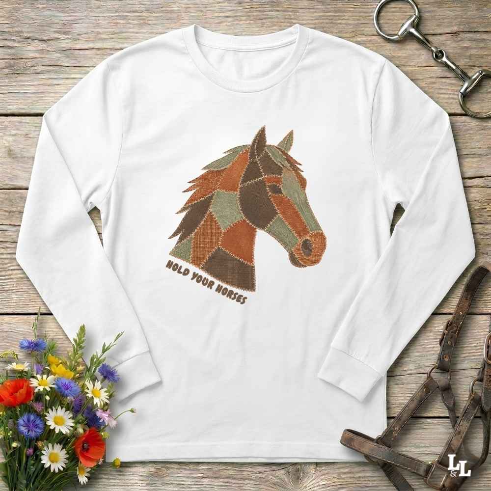 A white long-sleeve t-shirt with a graphic of a horse head made of patchwork shapes in brown, green, and orange, with the text 'HOLD YOUR HORSES' printed below it. The shirt is displayed flat on a rustic wooden background with equestrian-themed props including a halter, leather bridle, and a small bouquet of wildflowers.
