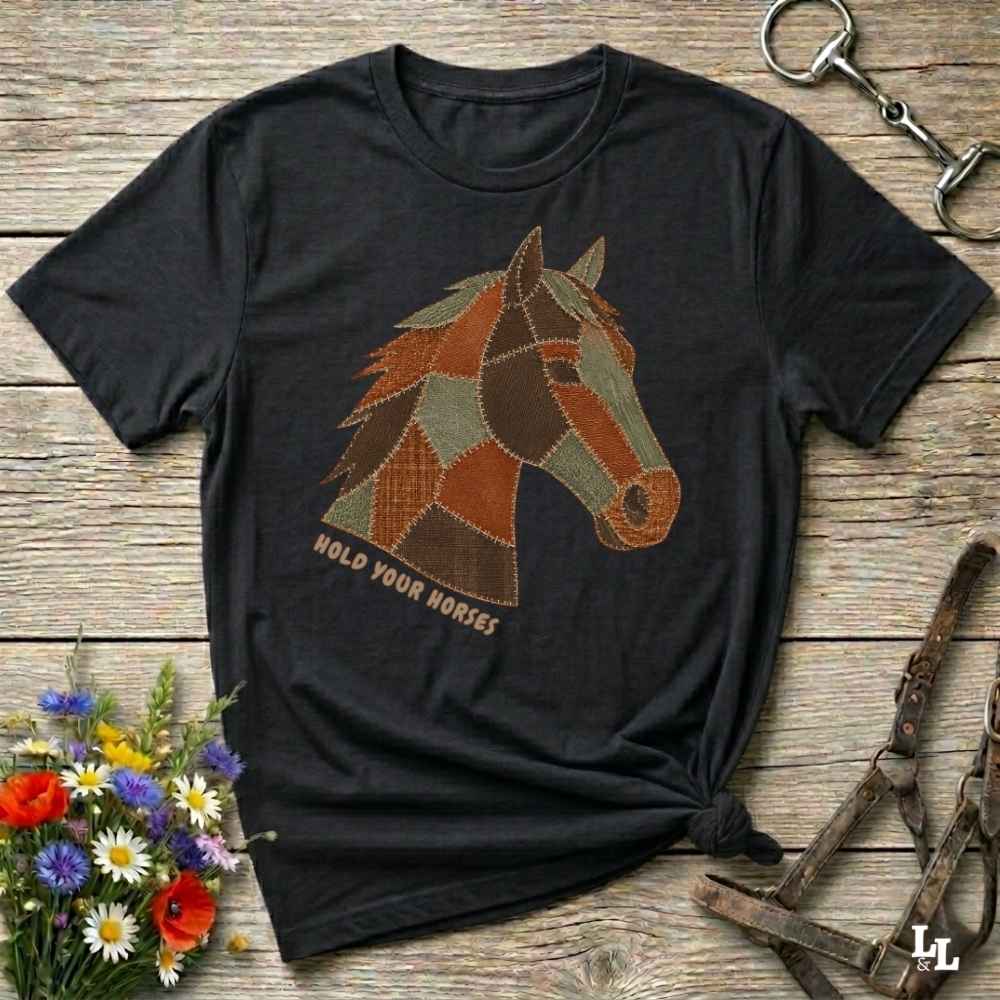 A black short-sleeve graphic T-shirt laid flat on a rustic wooden surface, featuring a patchwork horse head design in brown, tan, and muted green tones with the text 'HOLD YOUR HORSES' beneath it. Decorative flowers and equestrian hardware are placed around the shirt.