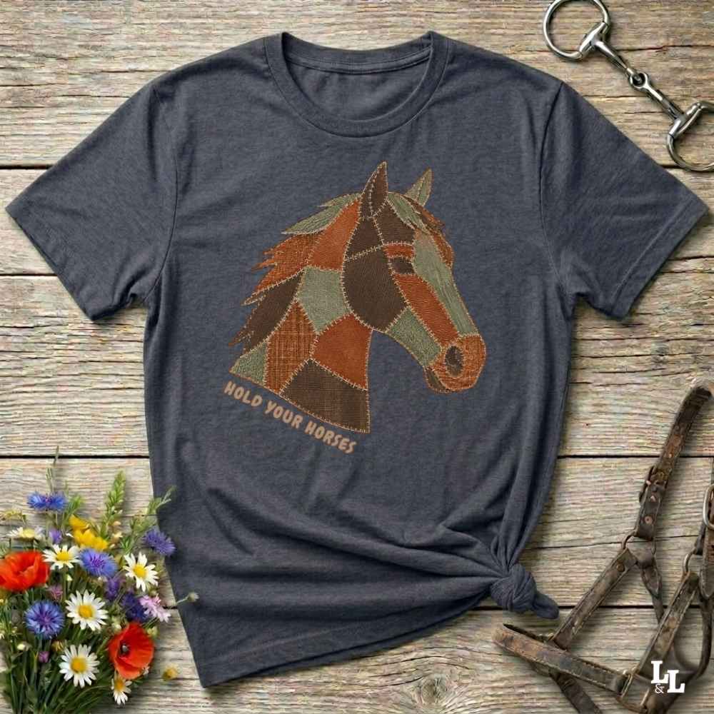 A dark heather gray t-shirt with a patchwork horse graphic and the text 'HOLD YOUR HORSES' printed on the front, styled on a rustic wooden background with equestrian accessories and flowers.