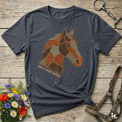 A dark heather gray t-shirt with a patchwork horse graphic and the text 'HOLD YOUR HORSES' printed on the front, styled on a rustic wooden background with equestrian accessories and flowers.