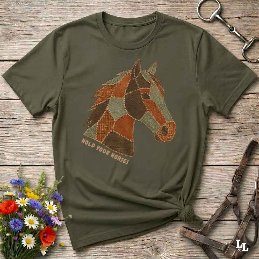 A green short-sleeve graphic T-shirt laid flat on a rustic wooden surface, featuring a patchwork horse head design and the words 'HOLD YOUR HORSES.' Decorative flowers, a halter, and a metal tack piece surround the shirt.