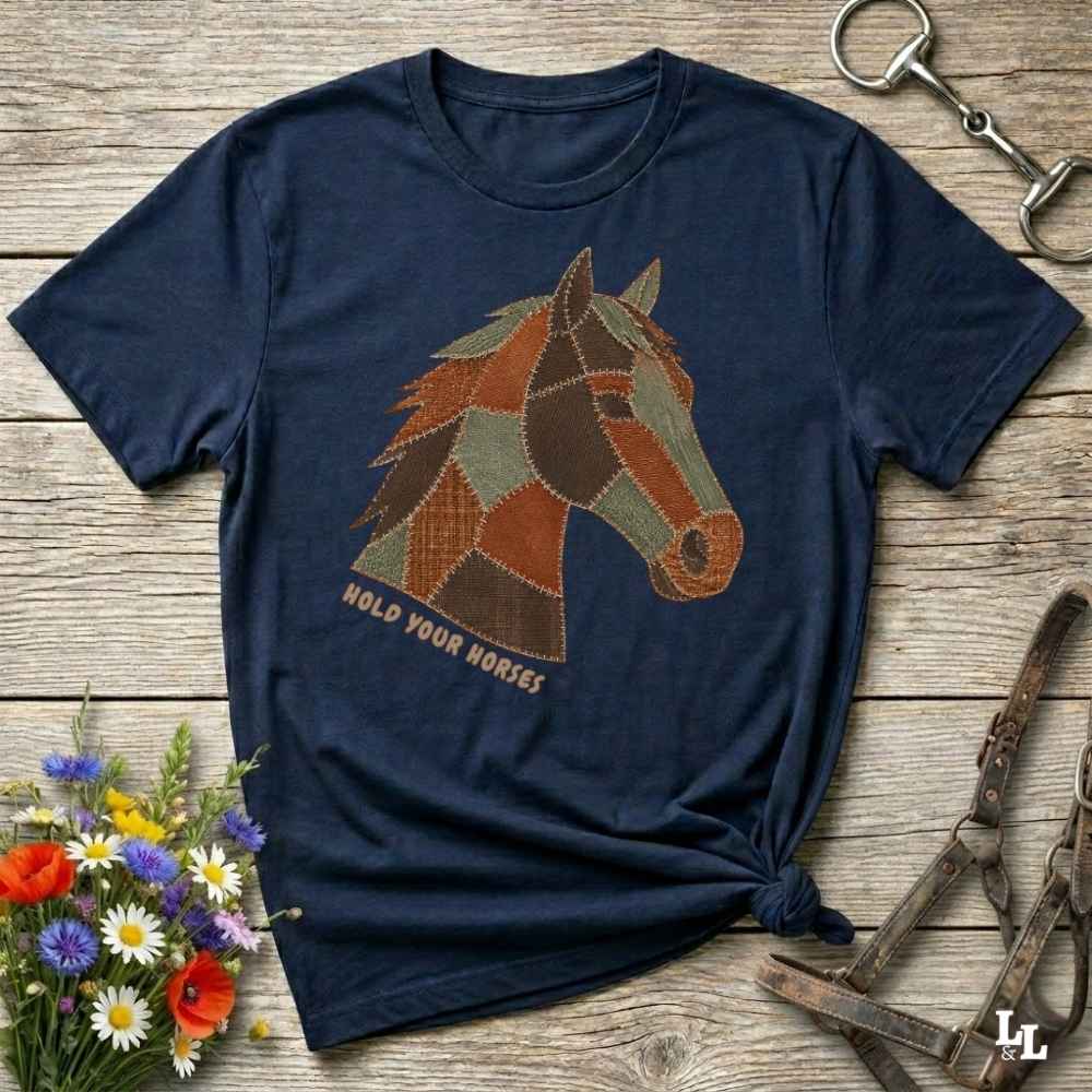A navy blue short-sleeve graphic T-shirt laid flat on a rustic wooden surface, featuring a patchwork horse head design and the words 'HOLD YOUR HORSES.' Decorative flowers, a halter, and a metal tack piece surround the shirt.