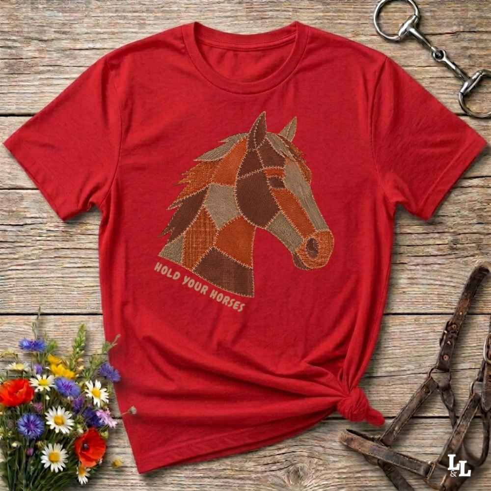 A red short-sleeve graphic T-shirt laid flat on a rustic wooden surface, featuring a patchwork horse head design and the words 'HOLD YOUR HORSES.' Decorative flowers, a halter, and a metal tack piece surround the shirt.