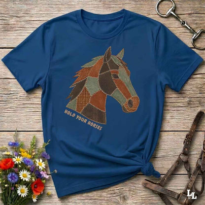 A blue short-sleeve graphic T-shirt laid flat on a rustic wooden surface, featuring a patchwork-style horse head design with the words 'HOLD YOUR HORSES' beneath it. Decorative flowers and equestrian straps are placed around the shirt.