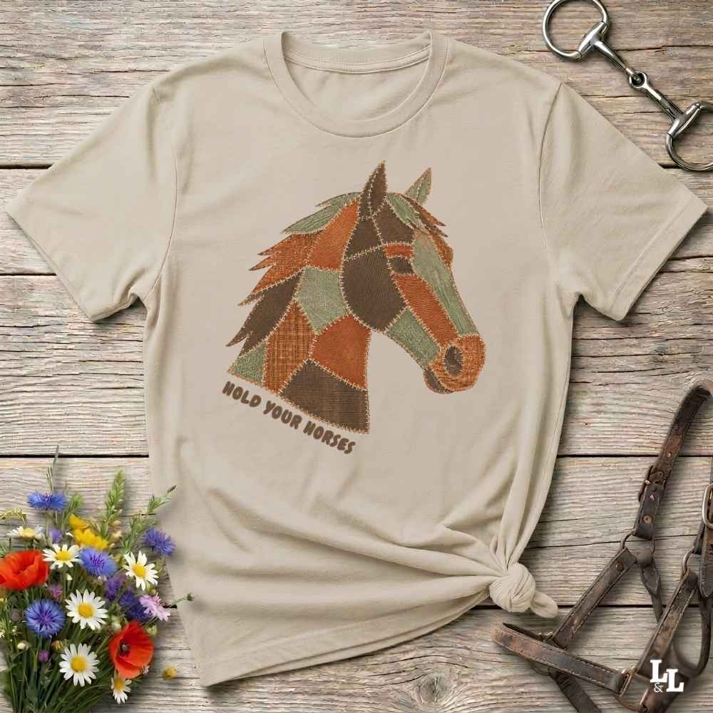 A beige short-sleeve t-shirt with a graphic of a horse head made of patchwork shapes in brown, orange, and green, with the text 'HOLD YOUR HORSES' underneath. The shirt is styled in a flat-lay on a rustic wooden background with equestrian accessories and a small bouquet of wildflowers.