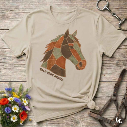 A beige short-sleeve t-shirt with a graphic of a horse head made of patchwork shapes in brown, orange, and green, with the text 'HOLD YOUR HORSES' underneath. The shirt is styled in a flat-lay on a rustic wooden background with equestrian accessories and a small bouquet of wildflowers.