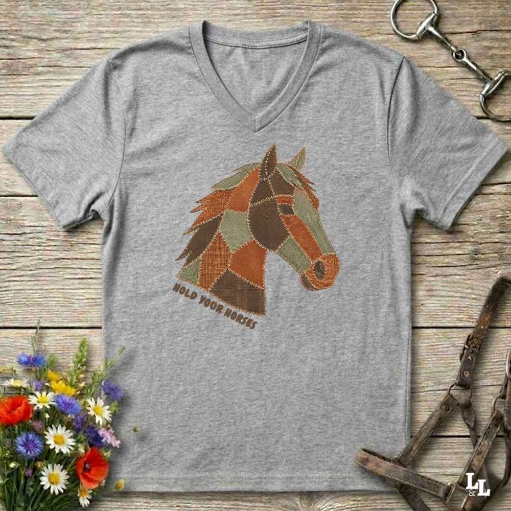 A grey V-neck t-shirt laid flat on a rustic wooden surface, featuring a stylized patchwork horse head graphic with the text 'HOLD YOUR HORSES' underneath. The shirt is styled with equestrian tack, a small bouquet of wildflowers, and a metal horse bit.